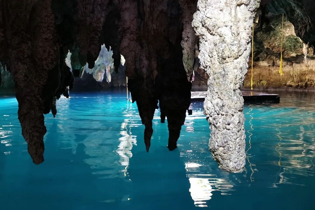 Des stalactites pendent du plafond de la grotte au-dessus de l'eau bleue claire, reflétant les formations rocheuses et la lumière de l'entrée de la grotte.
