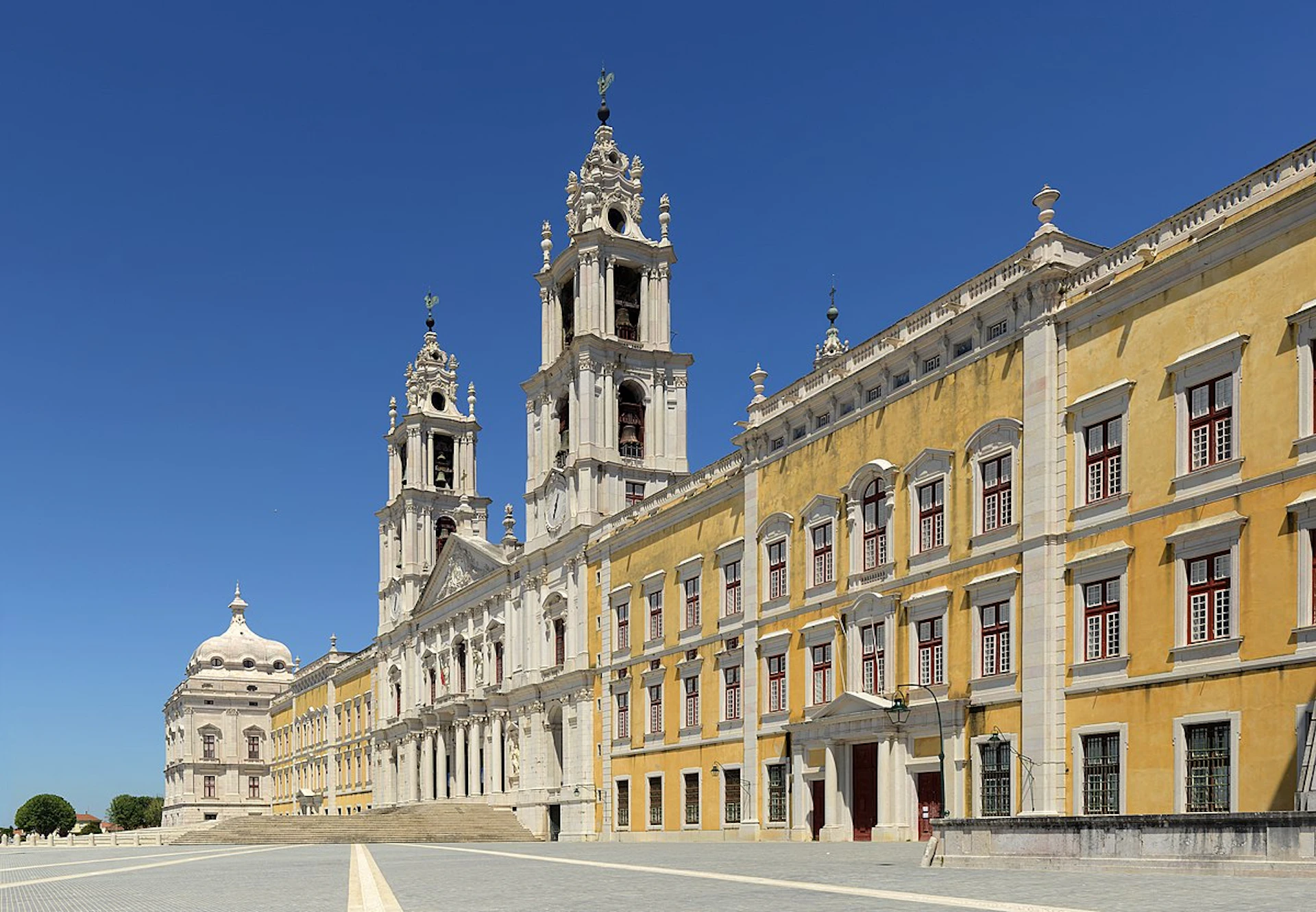 A large yellow and white baroque building with two tall towers and numerous windows under a clear blue sky.