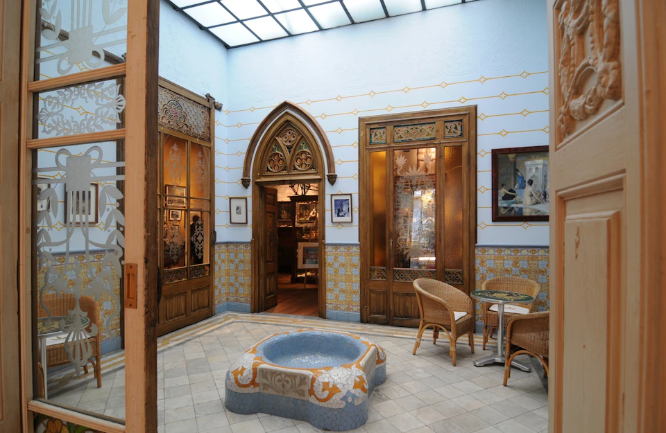 Ornate indoor courtyard with wooden doors, tiled walls, wicker furniture, a round stone fountain, and a glass ceiling.
