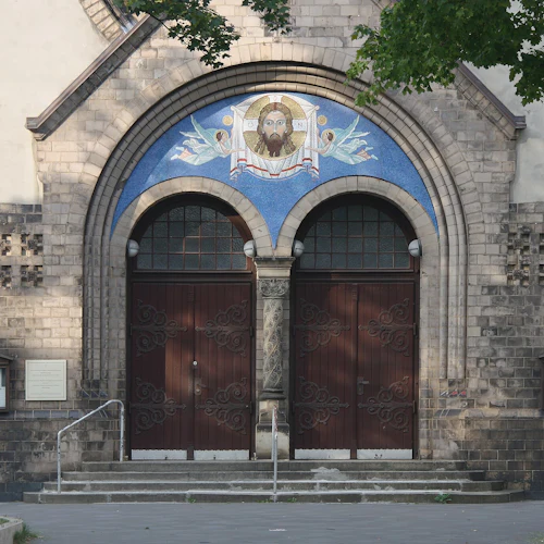 Tegelstensfasad med två utsmyckade trädörrar under en mosaik av en skäggig figur. Anslagstavlor flankerar trappan.