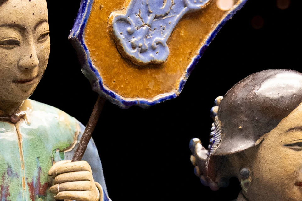 Two decorative ceramic figures in traditional attire, one holding a shield-like object, against a black background.