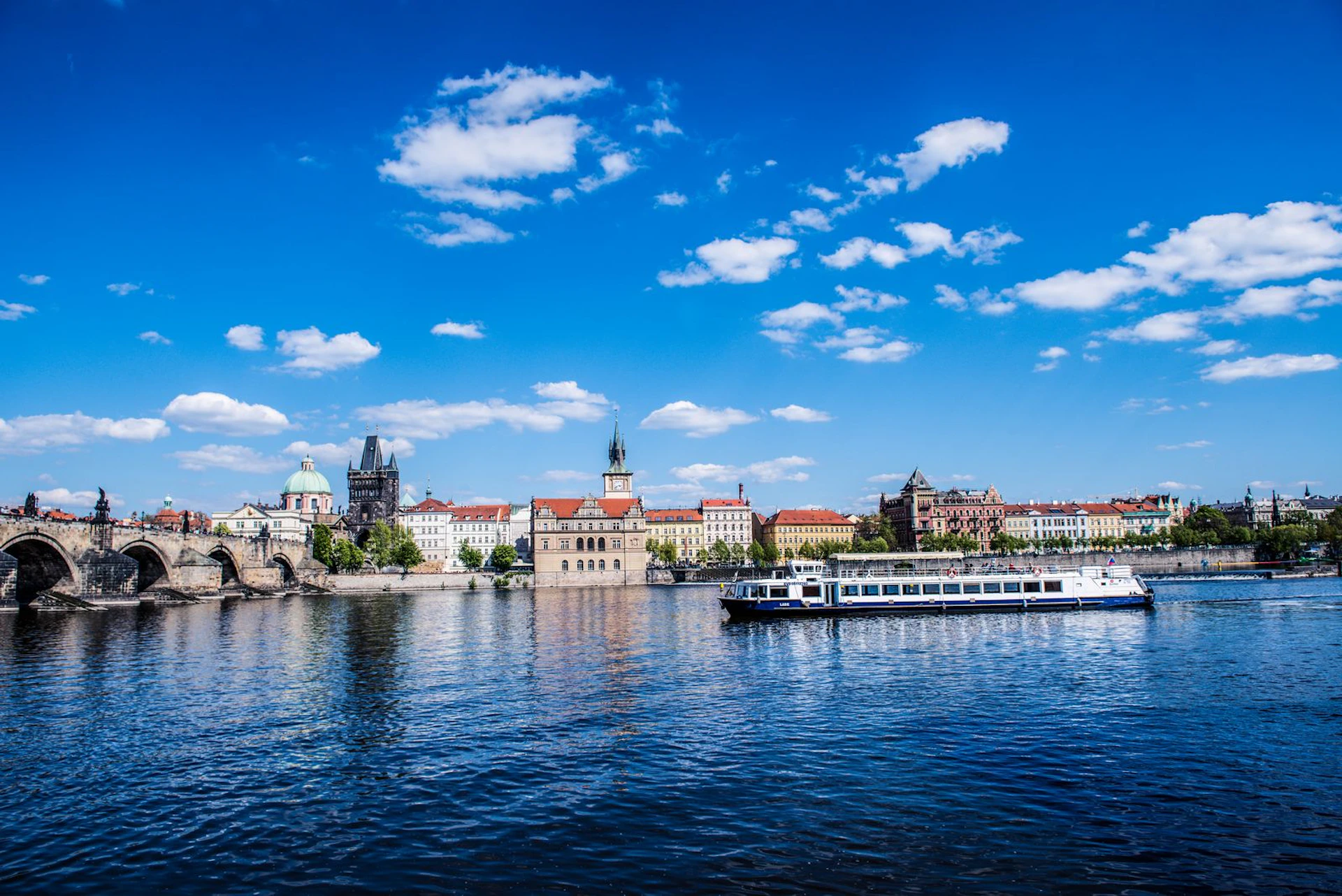 A boat sails on a river with historical buildings, including a tower and church, in the background under a bright blue sky with clouds.