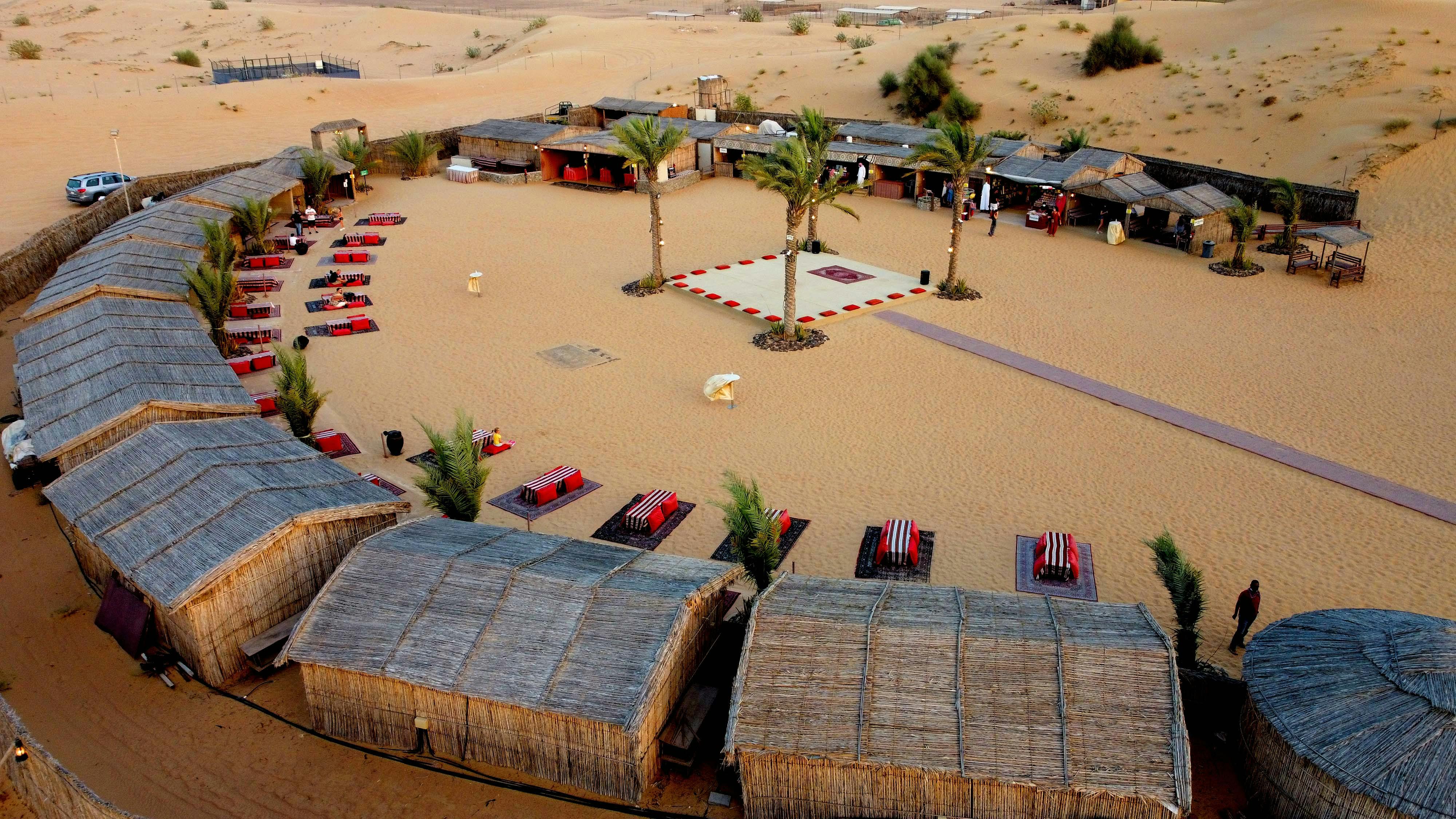Desert campsite with woven huts, red and black cushioned seating, palm trees, and a central square with a decorative rug.