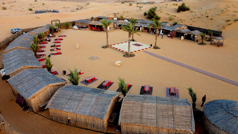 Desert campsite with woven huts, red and black cushioned seating, palm trees, and a central square with a decorative rug.