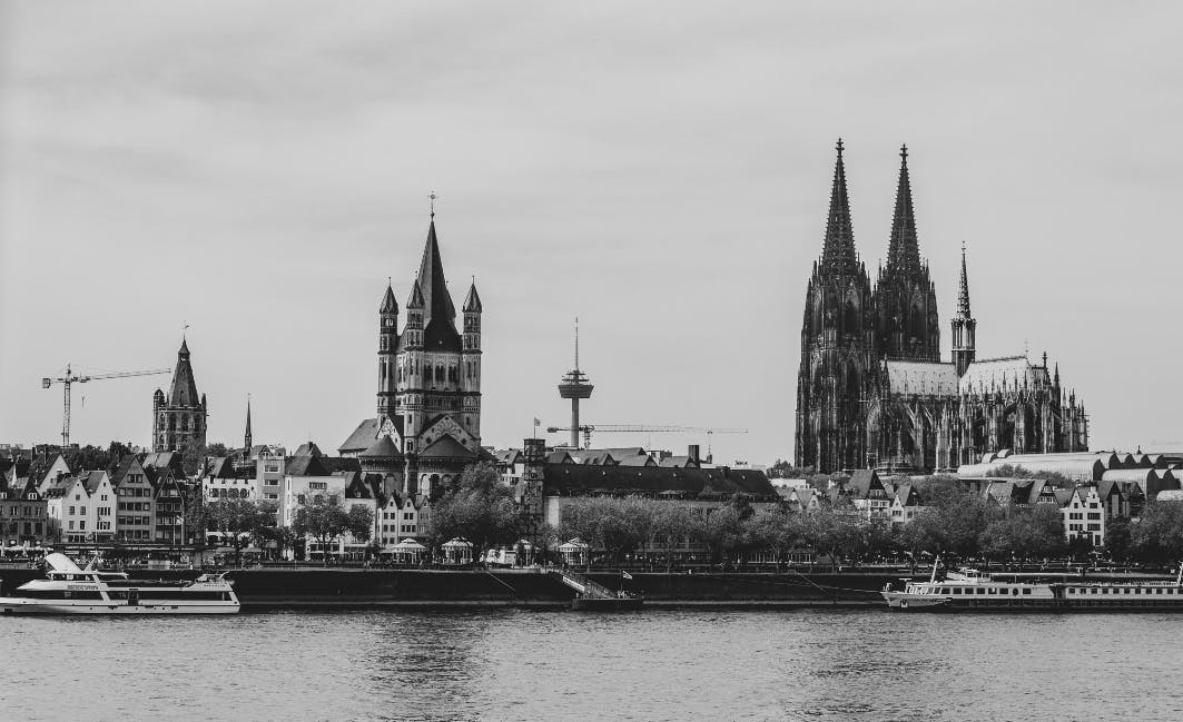 City skyline with historic churches, modern tower, and cranes. Boats on the river in the foreground. Black and white photograph.