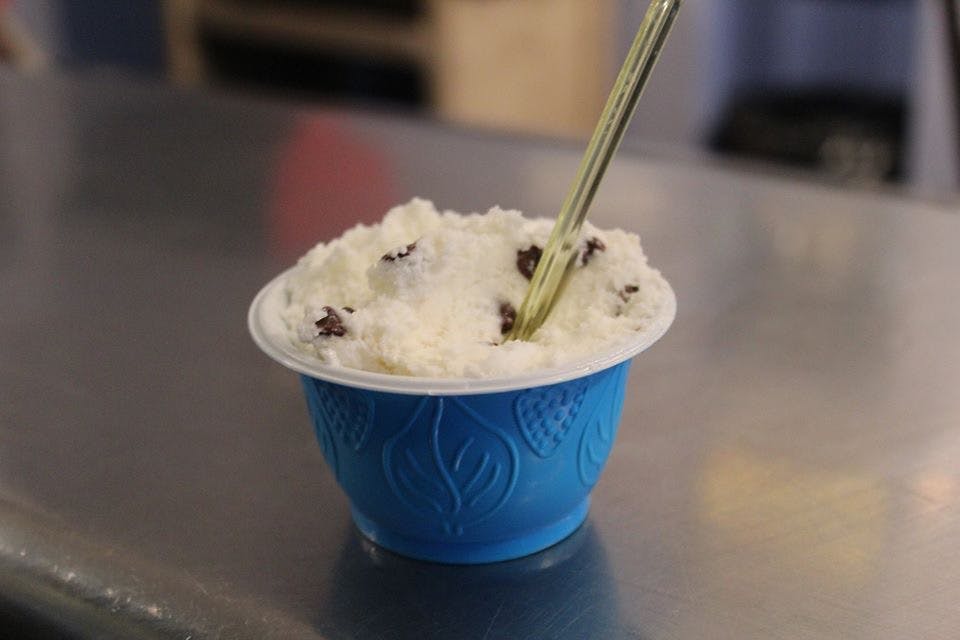 Una coppa blu riempita di gelato bianco con gocce di cioccolato, posizionata su una superficie riflettente, con un cucchiaio inserito.