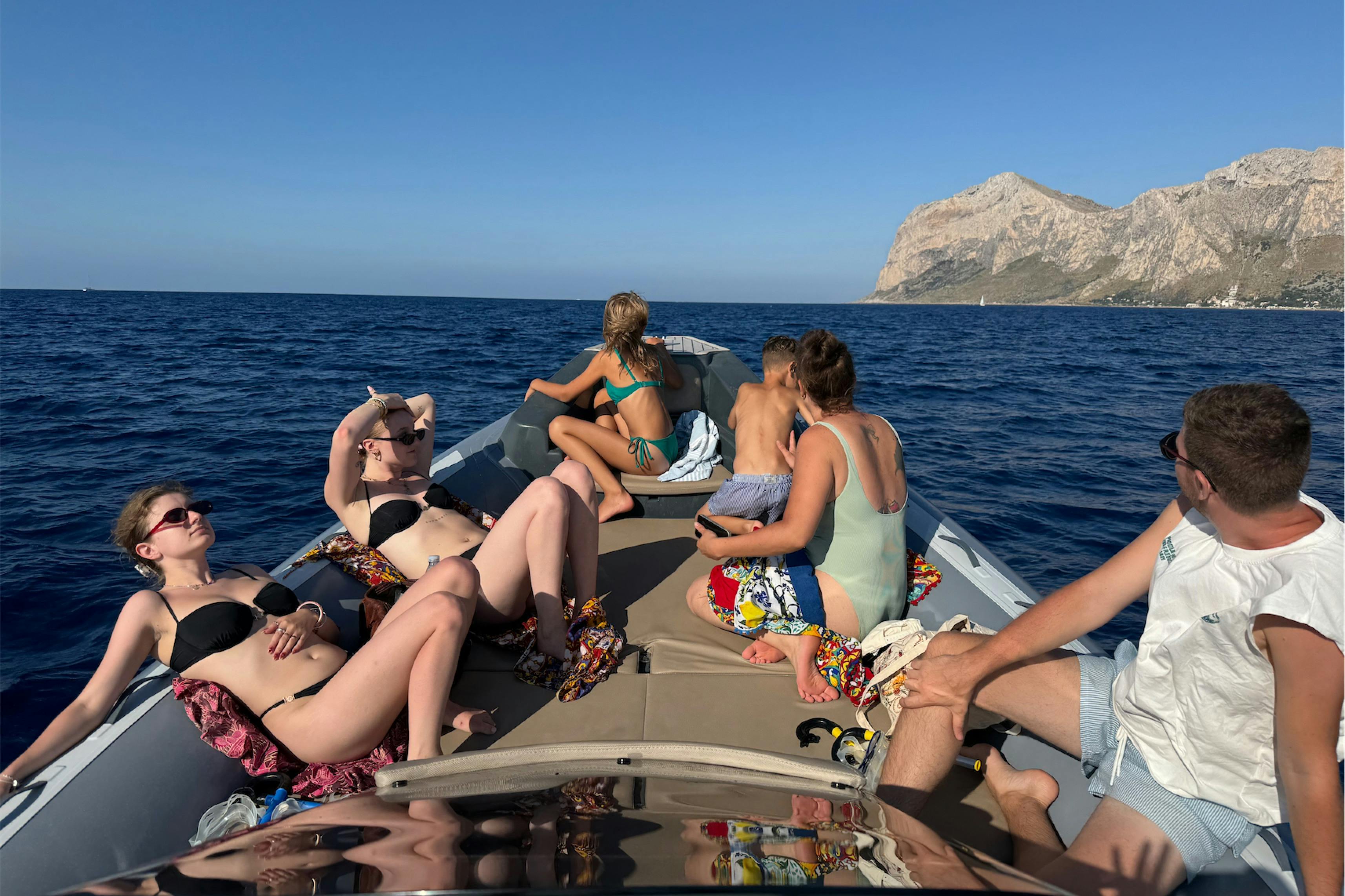 Persone che prendono il sole e si rilassano su una barca in mare aperto. Una costa rocciosa è visibile sullo sfondo sotto un cielo limpido.