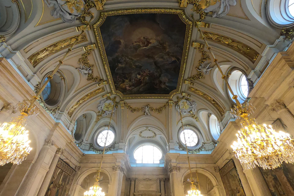 Ornate interior of a hall with a painted ceiling, large arched windows, and golden chandeliers.