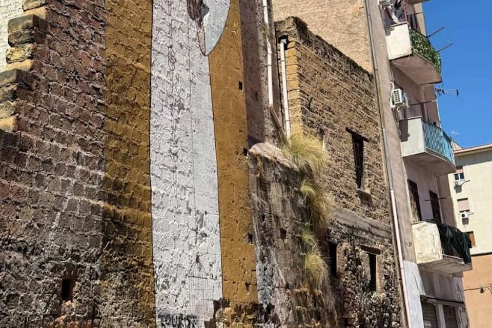 Old stone building with a partial mural on the wall and grass growing from a crevice. Visible rooftops and a window with bars.