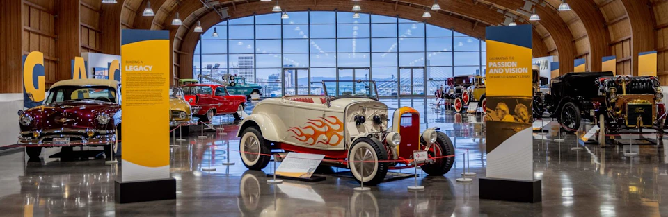 Vintage car with a flame decal on display in a large, windowed showroom. Other classic cars are visible in the background.
