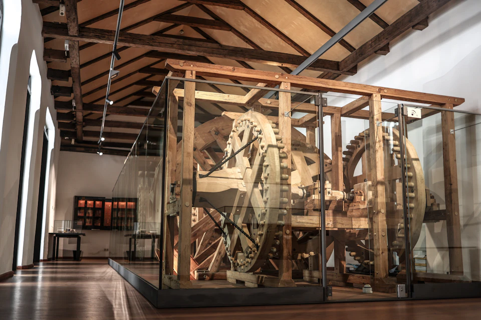 A large wooden mechanical structure displayed behind glass in a museum with a high wooden-beamed ceiling.