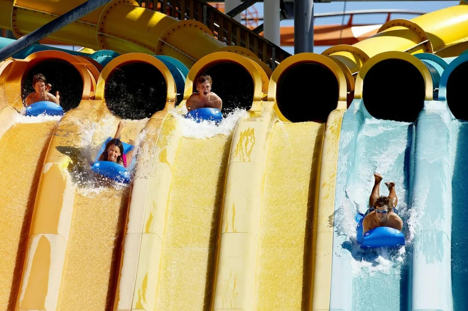 Tres persones lliscant per tobogans d'aigua paral·lels, cadascun en un tub inflable, amb esquitxades d'aigua al seu voltant.