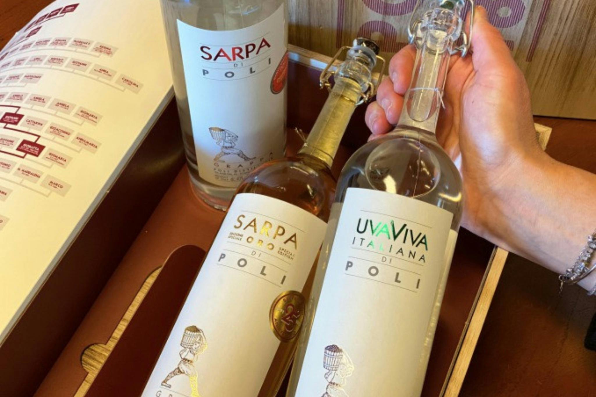 Τρία μπουκάλια Poli grappa βρίσκονται σε ένα ξύλινο κουτί, το ένα κρατιέται από το χέρι. Οι ετικέτες γράφουν "Sarpa di Poli", "Sarpa Oro" και "UvaViva Italiana".
