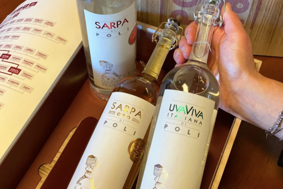 Three bottles of Poli grappa are in a wooden box, one held by a hand. Labels read "Sarpa di Poli," "Sarpa Oro," and "UvaViva Italiana."