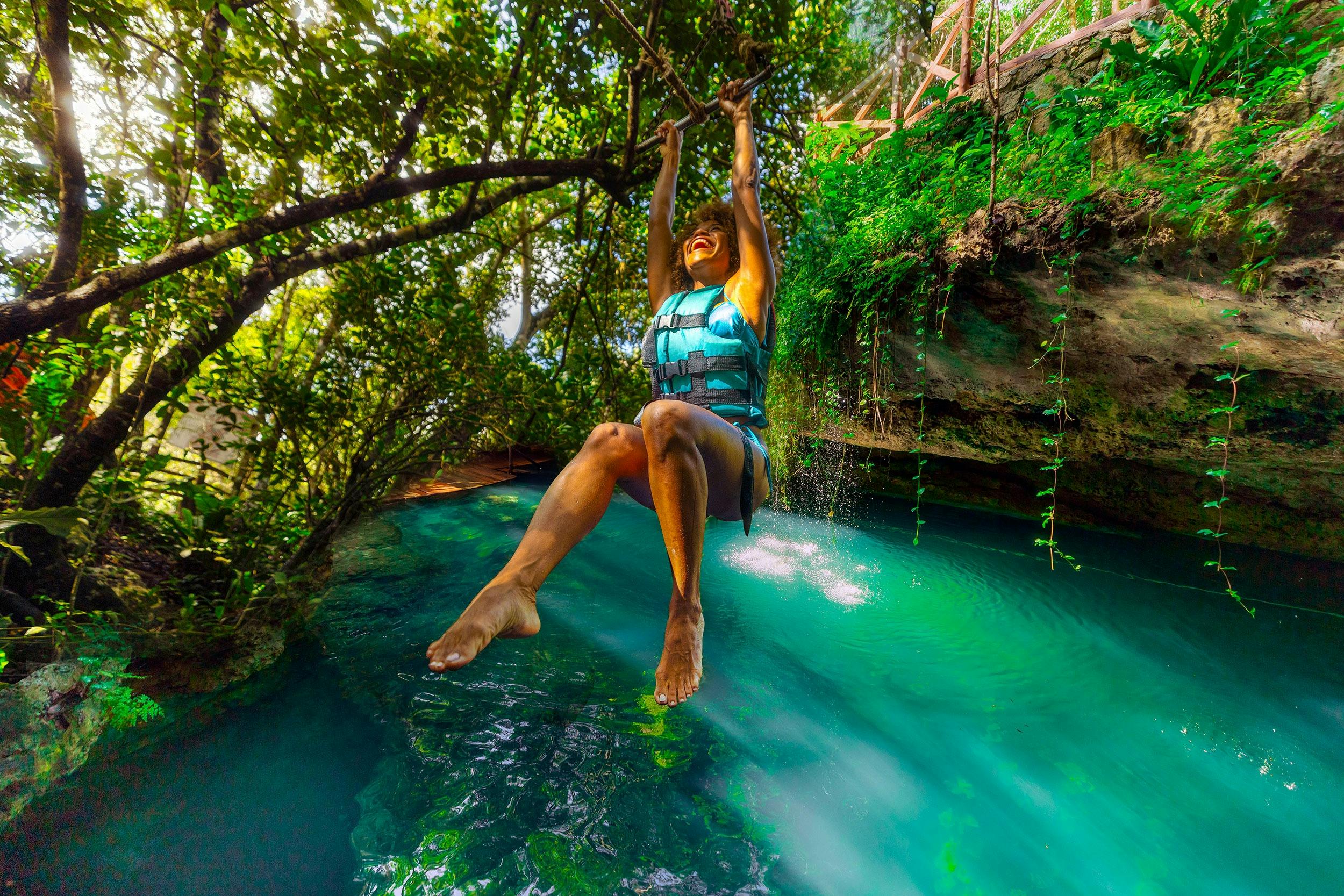 Una persona amb un xaleco salvavides es balanceja sobre una piscina d'aigua turquesa en un entorn forestal exuberant en un dia assolellat.