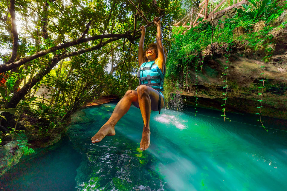 A person wearing a life vest swings over a turquoise water pool in a lush forest environment on a sunny day.
