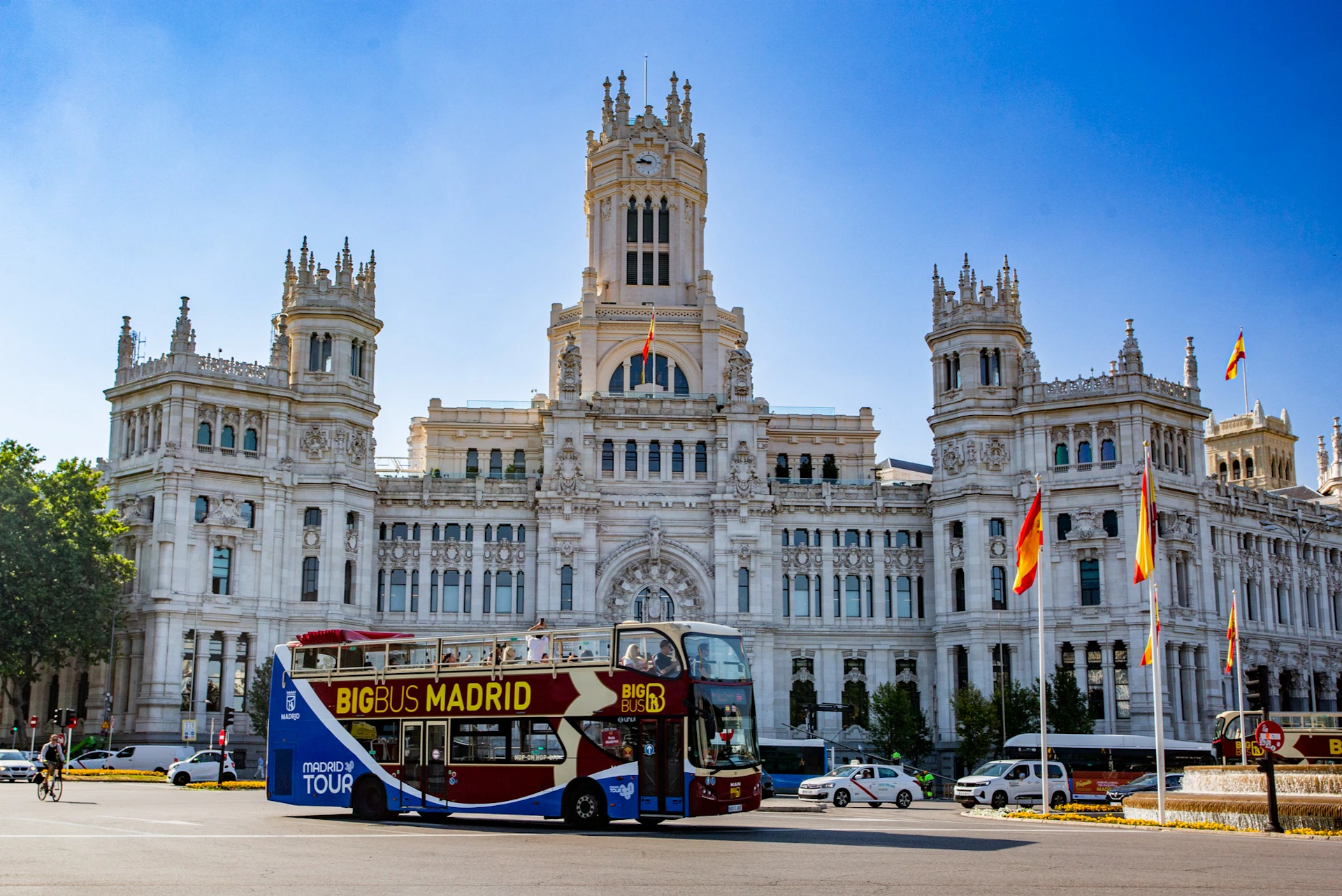 A red and yellow double-decker tour bus labeled 'Big Bus Madrid' in front of a large ornate building on a sunny day.