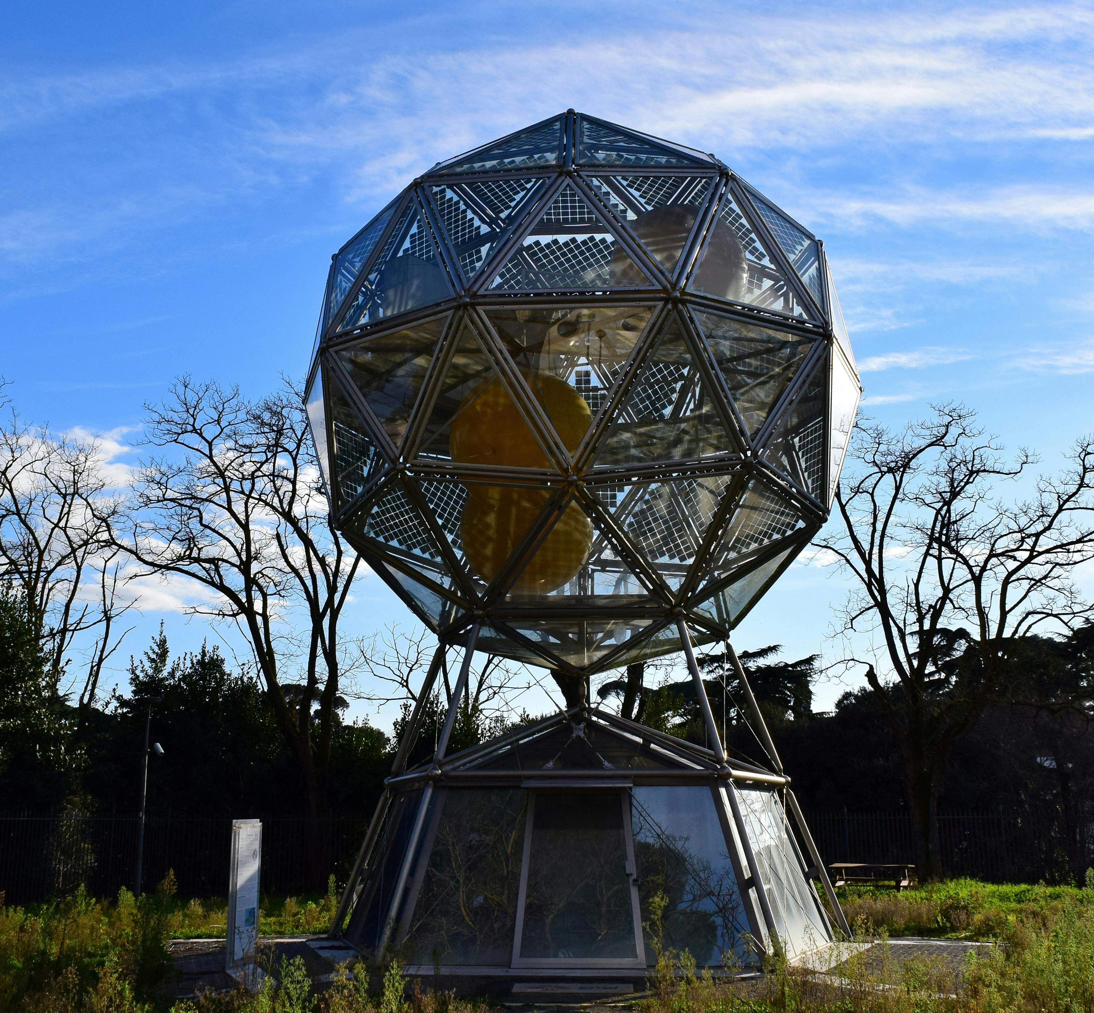Een geodetische koepel met glazen en metalen panelen, verhoogd op een voetstuk, omringd door bomen en gras onder een strakblauwe hemel.