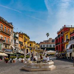Ett pulserande torg med färgglada byggnader, en central fontän, uteserveringar och en klarblå himmel.