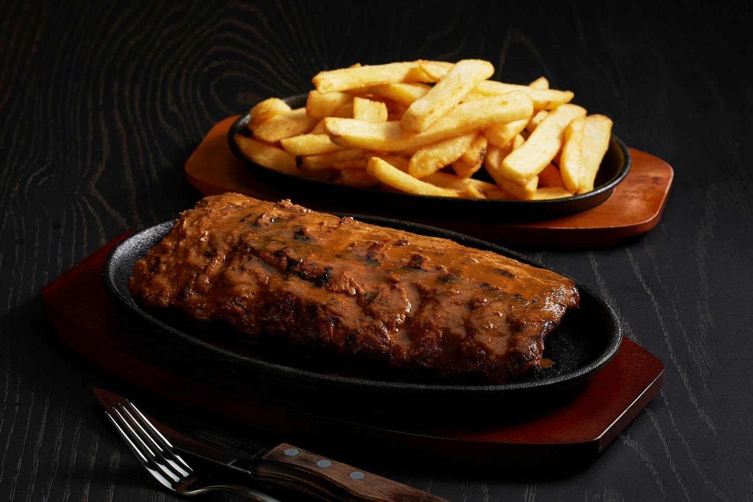Ein Teller mit saftigen Rippchen, serviert mit goldenen Pommes frites auf einer dunklen Oberfläche, begleitet von Gabel und Messer.