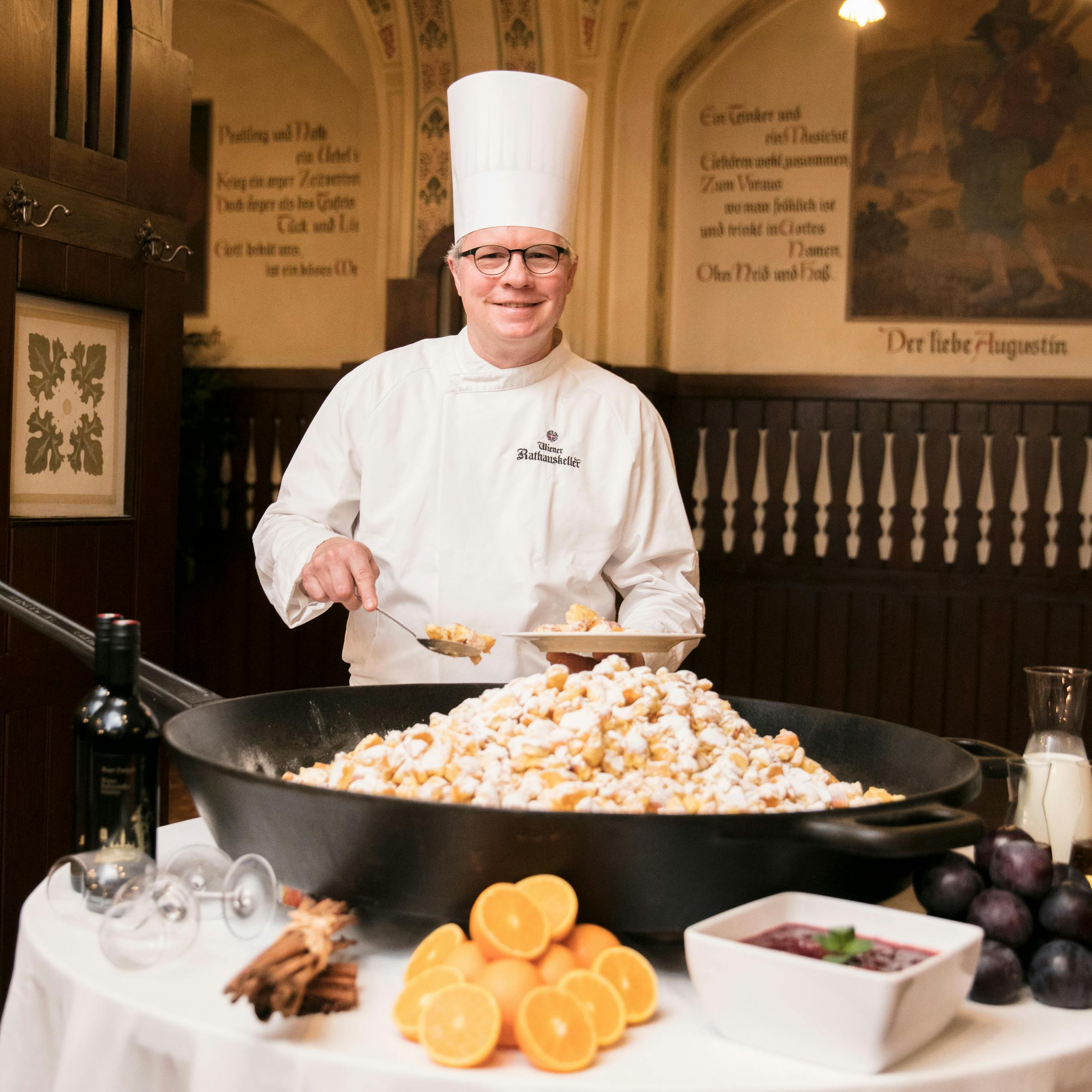 Un chef en uniforme et toque blanche prépare un grand plat avec du sucre en poudre, entouré d'oranges, de prunes, de bouteilles et d'un bol de sauce.