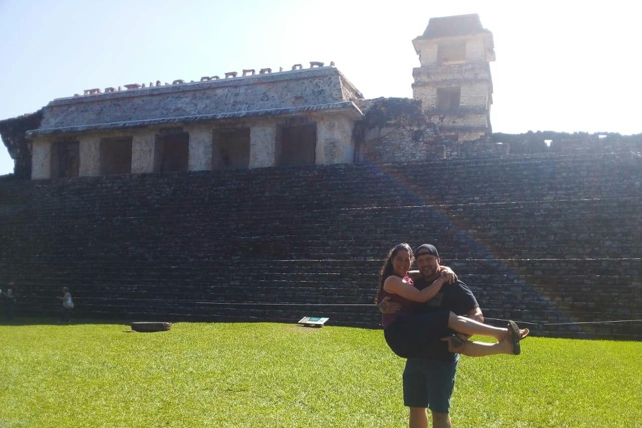A man holds a woman in his arms in front of an ancient stone structure with steps and a grassy foreground.