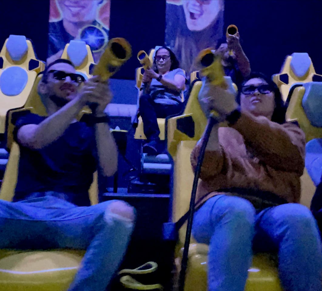 四个戴着3D眼镜的人坐在黄色座位上，瞄准橙色枪支，游乐园乘坐背景可见。