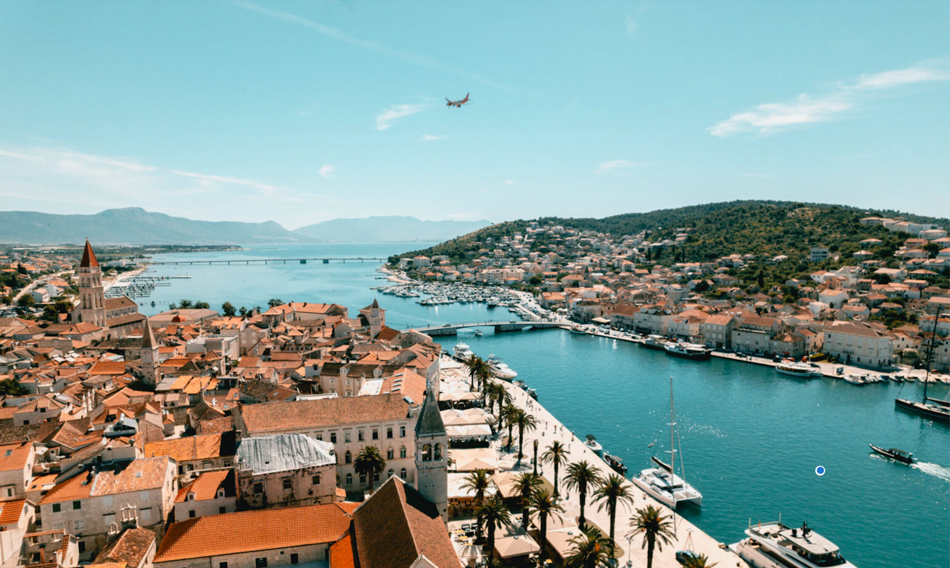 A coastal town with red-roofed buildings, a palm-lined waterfront, docked boats, and an airplane flying overhead.