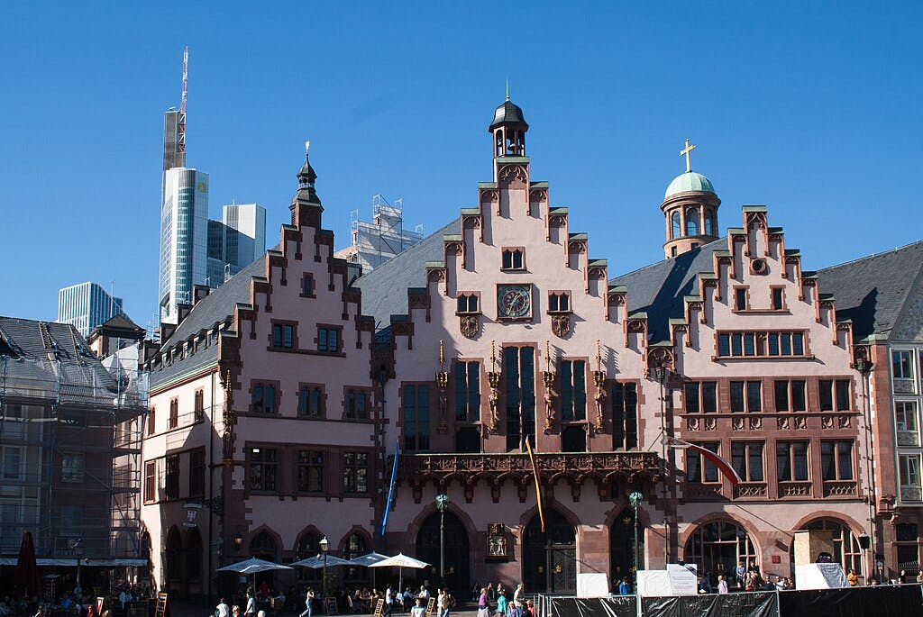 Ornate historic building with a stepped gable facade, surrounded by modern skyscrapers under a clear blue sky. People walk below.