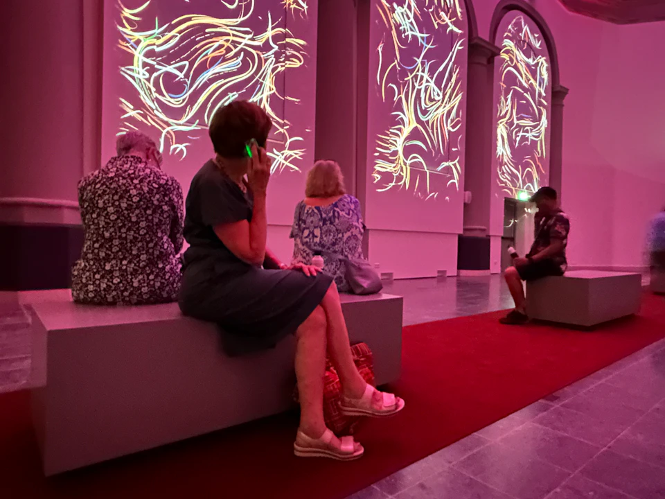 People sitting on benches in a dimly lit room with abstract light projections on the walls. One person is using a phone.