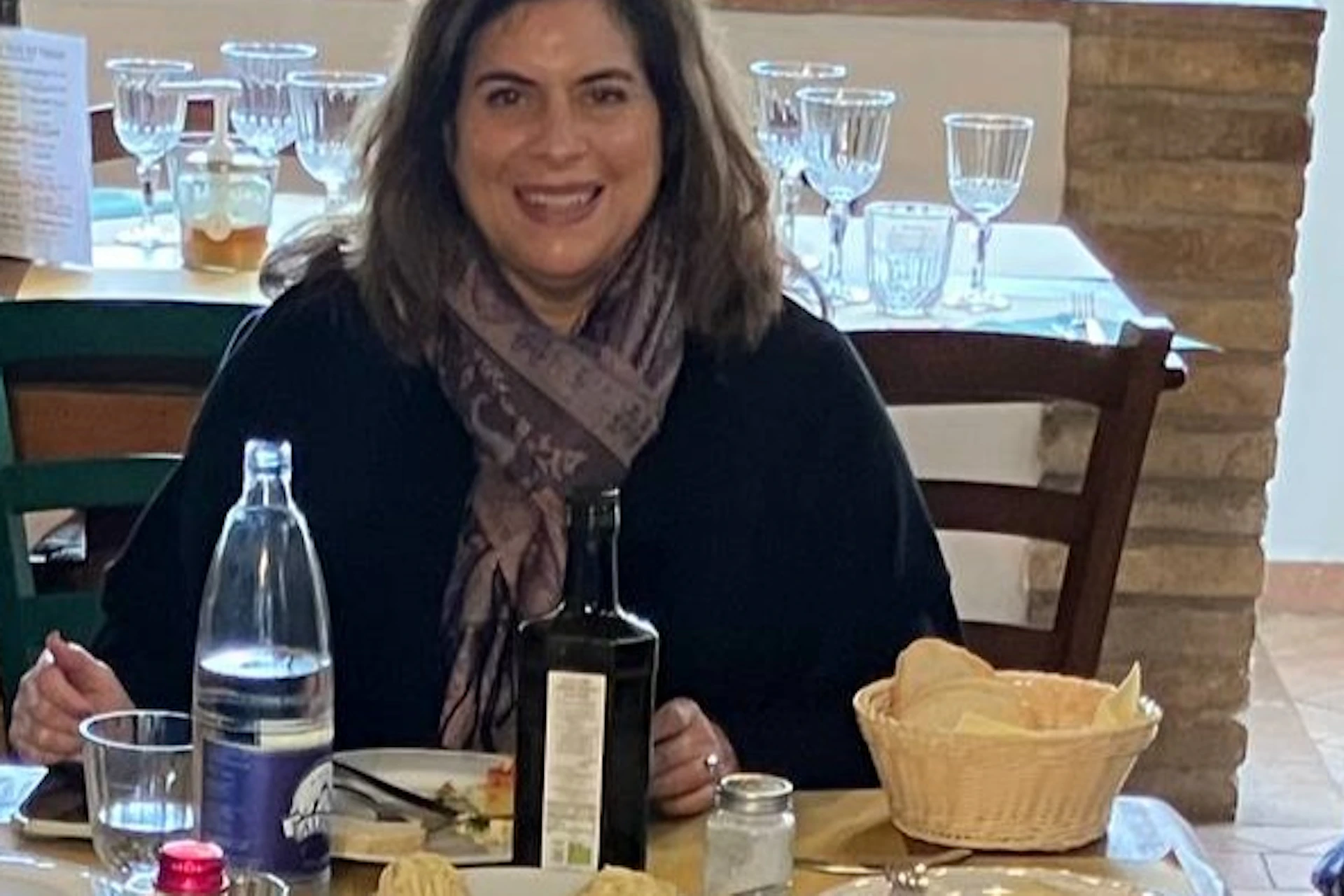 A group of six people seated at a table with plates of food, glasses, and bottles, smiling in a casual dining setting.