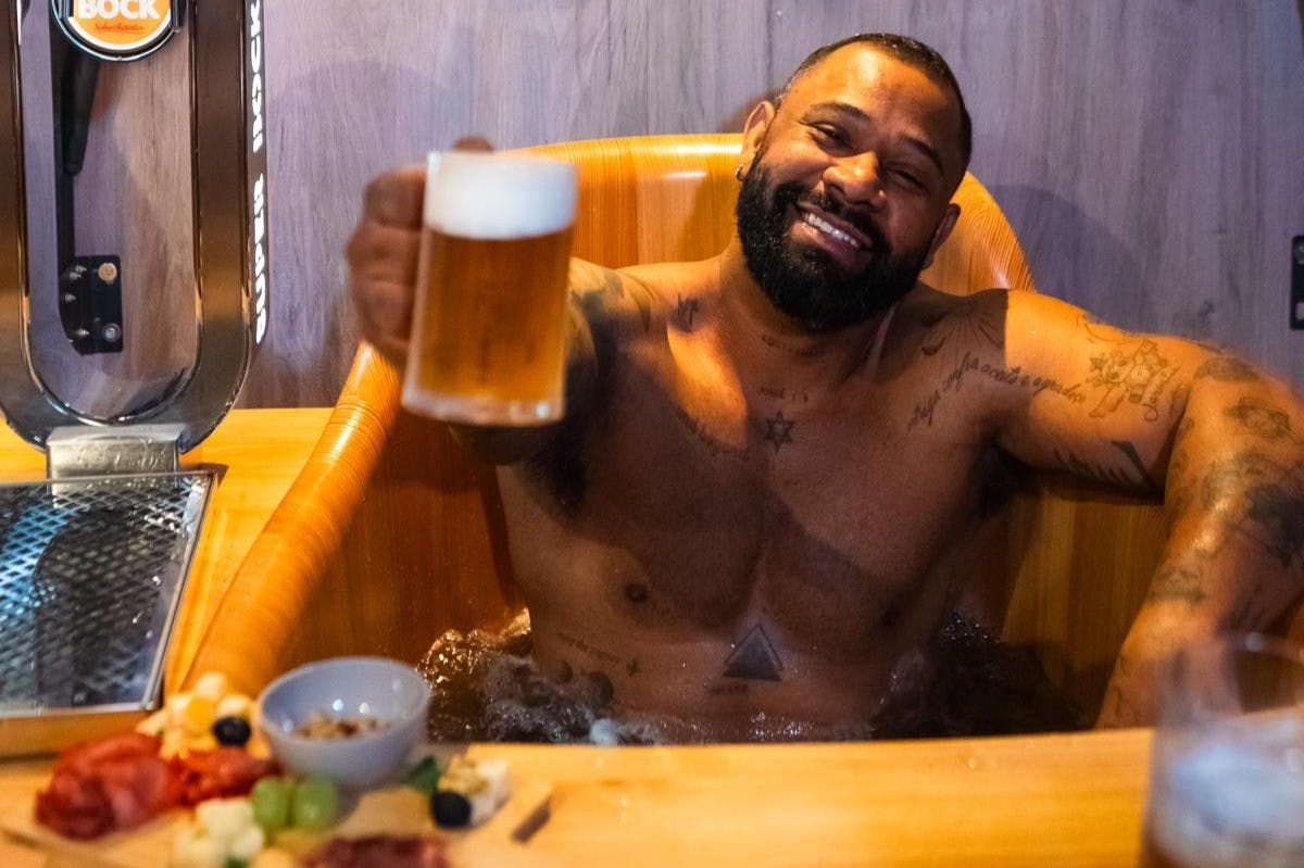 Ο άντρας με τα τατουάζ απολαμβάνει μια μπύρα χαλαρώνοντας σε ένα ξύλινο τζακούζι, χαμογελώντας και ανεβάζοντας το ποτό του.