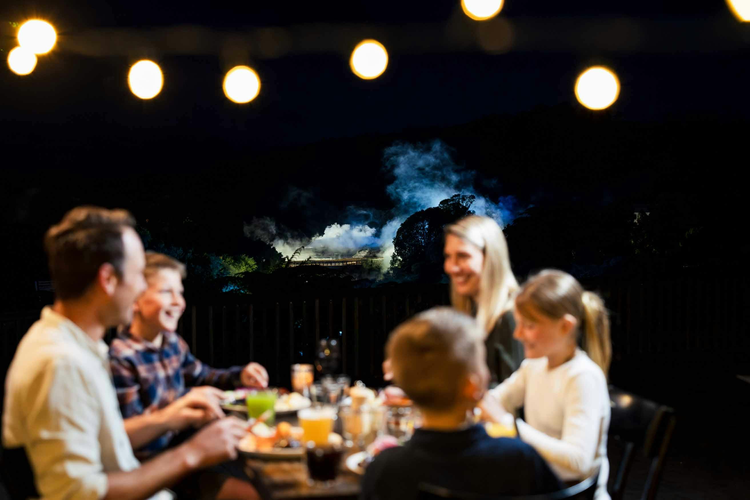 A group of people dining outdoors at night, with string lights above and a forested area with smoke visible in the background.
