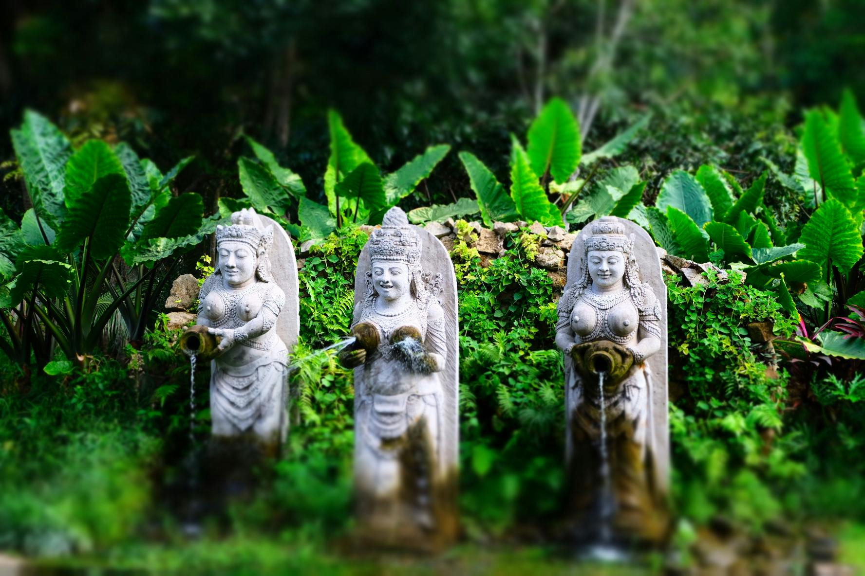 Three stone statues hold vases, with water flowing from them, surrounded by lush green foliage.