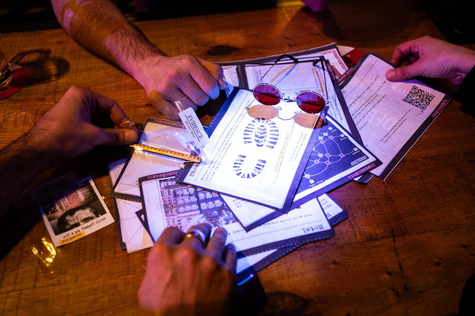 Hands holding various clue sheets, vial, and key; red sunglasses and papers with scientific diagrams on a wooden table.