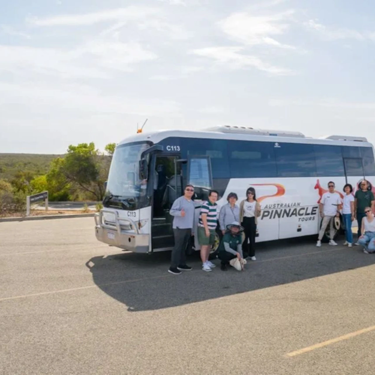 A group of people posing in front of a tour bus in a parking lot with a natural landscape in the background.