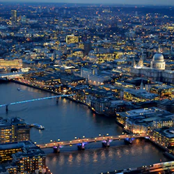 Flygfoto över en stad i skymningen med upplysta byggnader, broar över en flod och en framträdande kupolformad katedral.