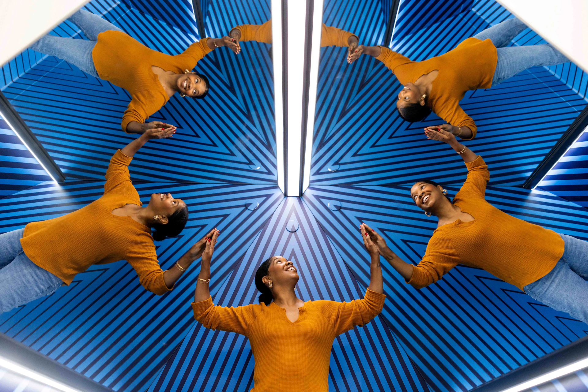 A person in an orange sweater is reflected multiple times against a blue, striped background with a central light.