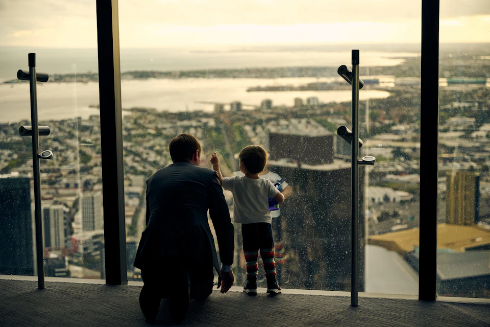 An adult and a child looking out through a high glass window at a cityscape with water and buildings in the distance.