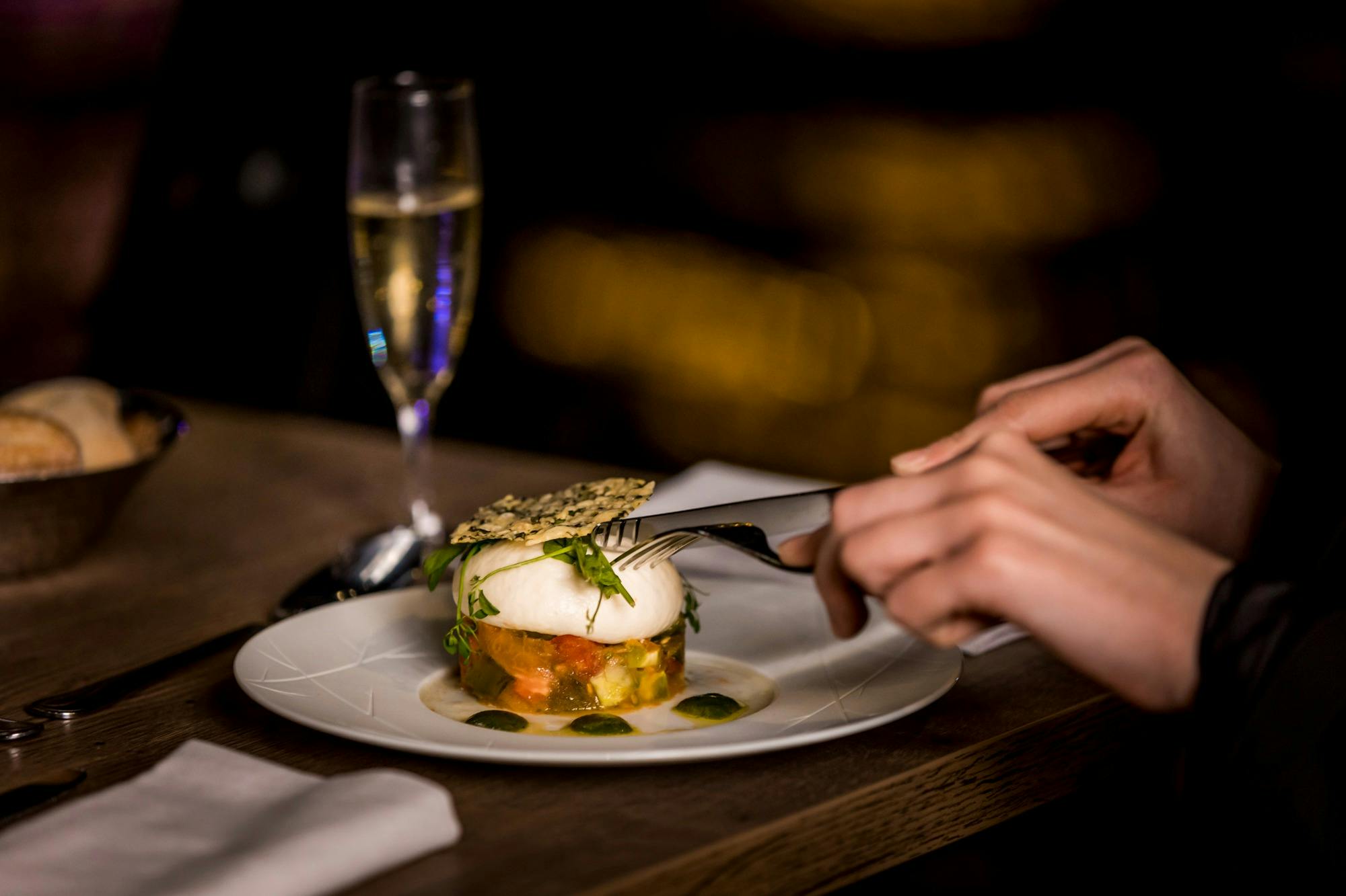 Eine Person, die mit Messer und Gabel ein Gourmetgericht auf einem weißen Teller isst, mit einem Glas Champagner im Hintergrund.