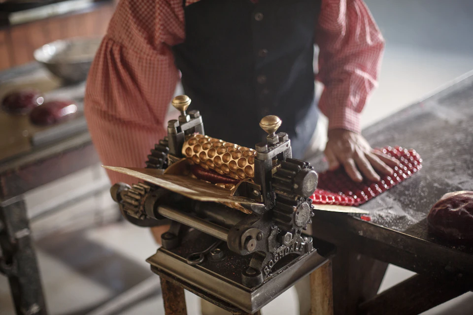 A person operates an old-fashioned, hand-cranked machine to press sheets of metal with a honeycomb pattern.