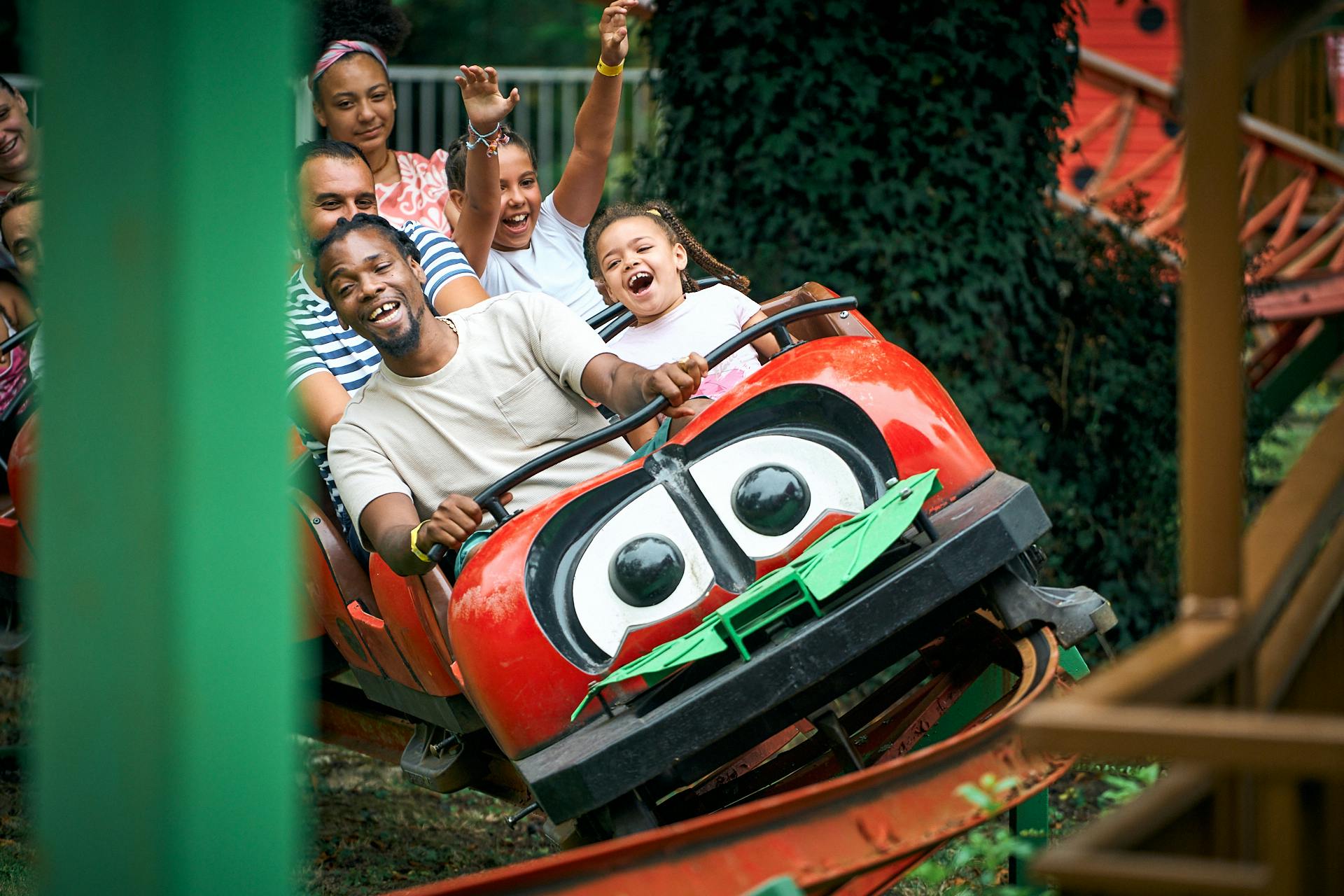 Skupina lidí se usmívá a zvedá ruce při jízdě na červené horské dráze s kreslenýma očima.