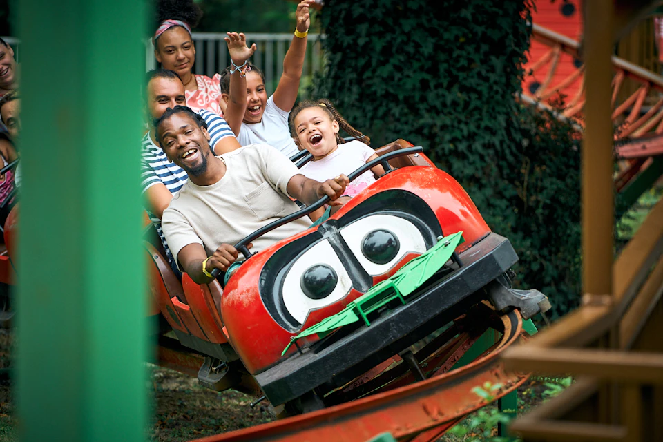 Group of people smiling and raising hands while riding a red rollercoaster car with cartoonish eyes.