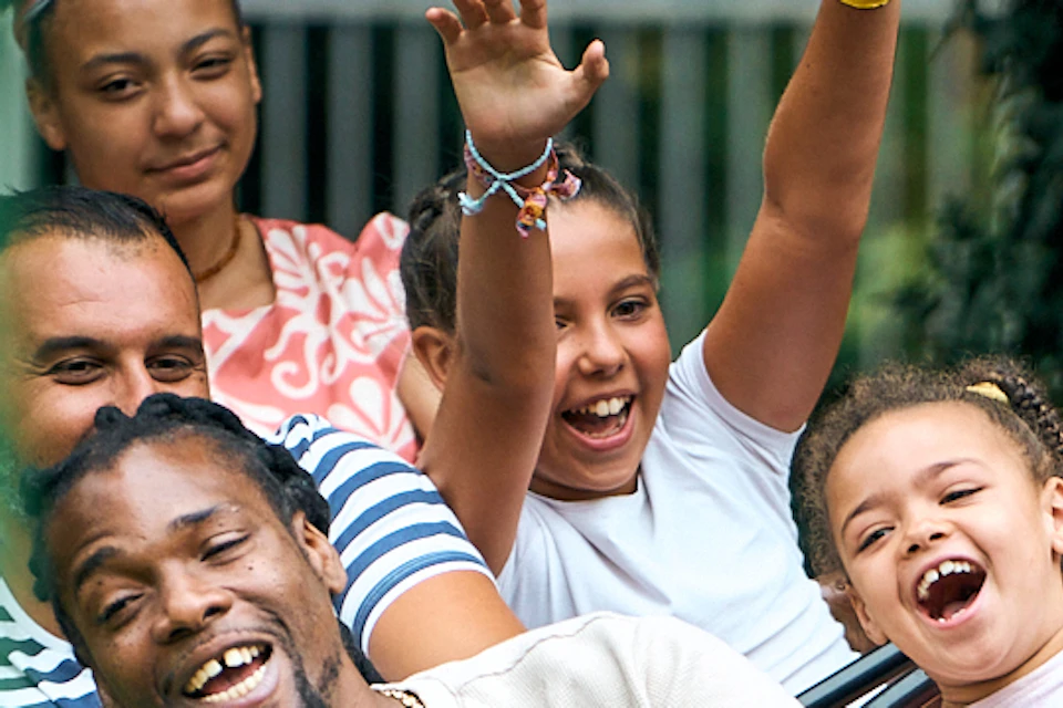 Group of people smiling and raising hands while riding a red rollercoaster car with cartoonish eyes.