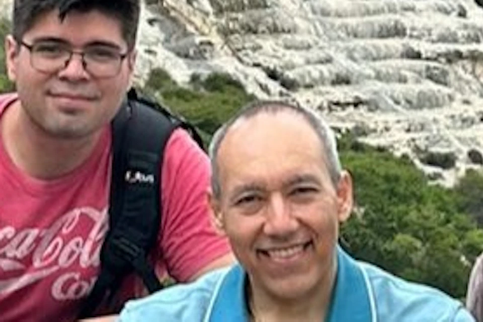 Four people posed in front of a scenic rock formation with a waterfall effect, surrounded by greenery and a cloudy sky.
