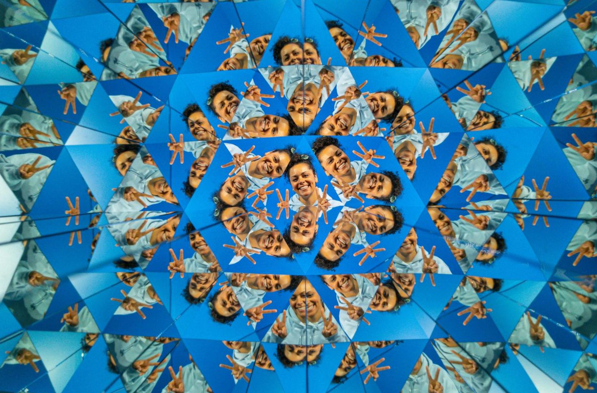 Ein kaleidoskopisches Muster mit einer lächelnden Person, die Friedenszeichen macht und sich vor einem blauen Hintergrund spiegelt.