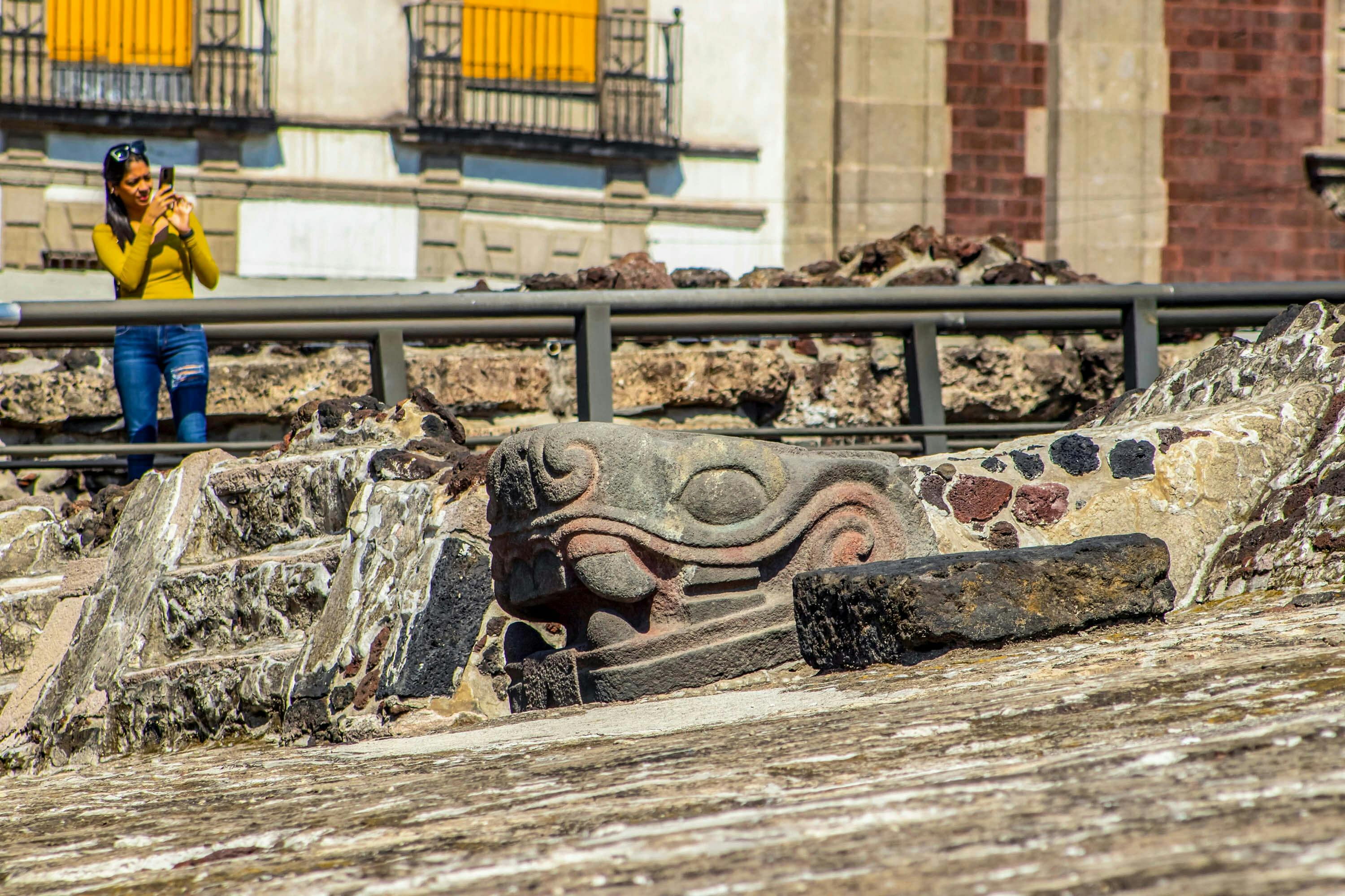 Scultura di serpente in pietra tra antiche rovine con parte di un edificio di epoca coloniale sullo sfondo.