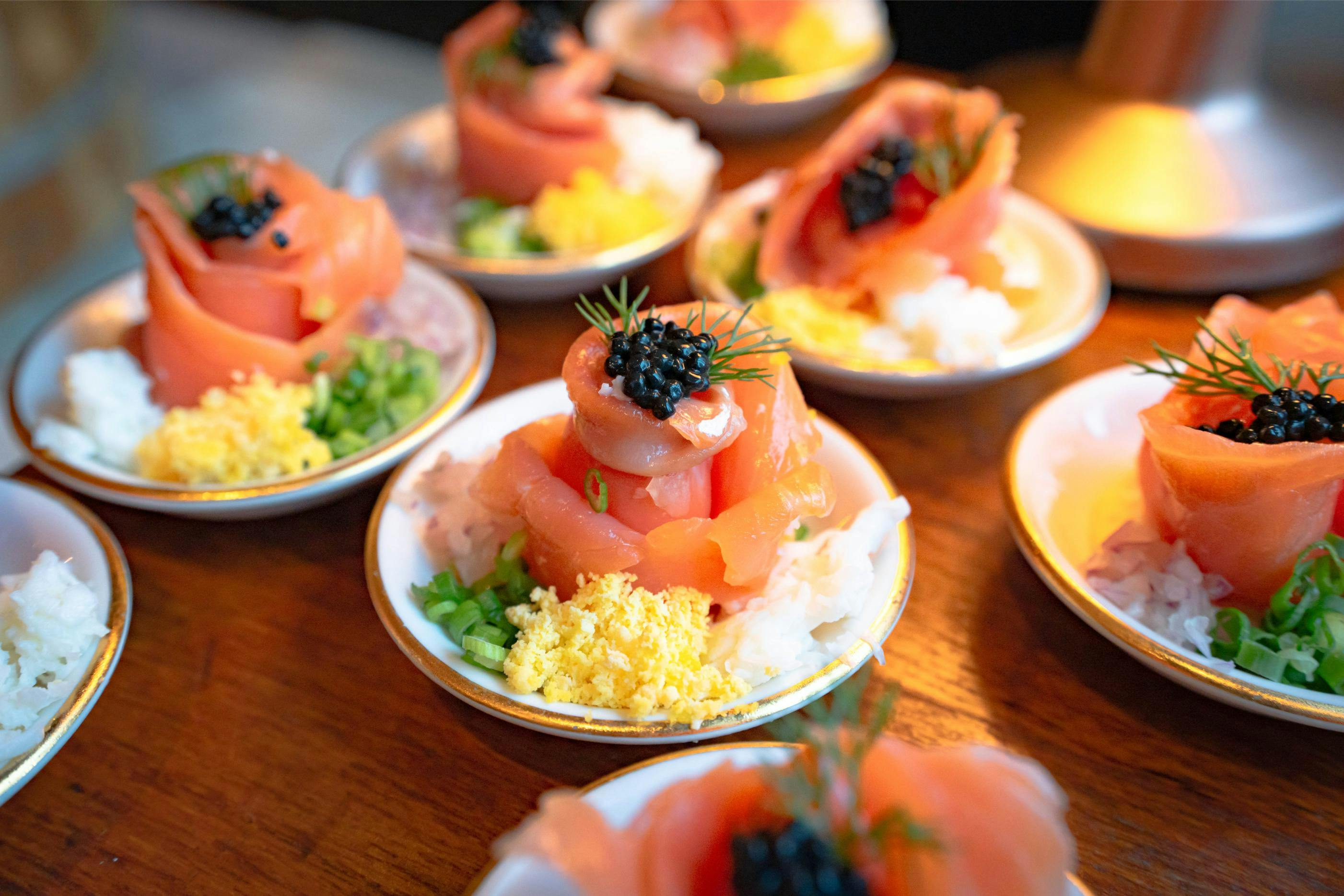 Plates of sashimi roses with black roe, garnished with herbs, served over white rice and surrounded by colorful sides.