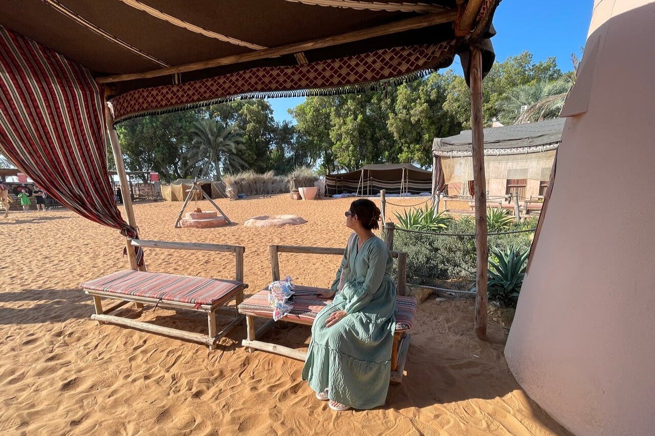 길고 연한 녹색 드레스를 입은 사람이 사막 캠프의 그늘진 공간 아래 벤치에 앉아 텐트와 식물이 근처에 있습니다.