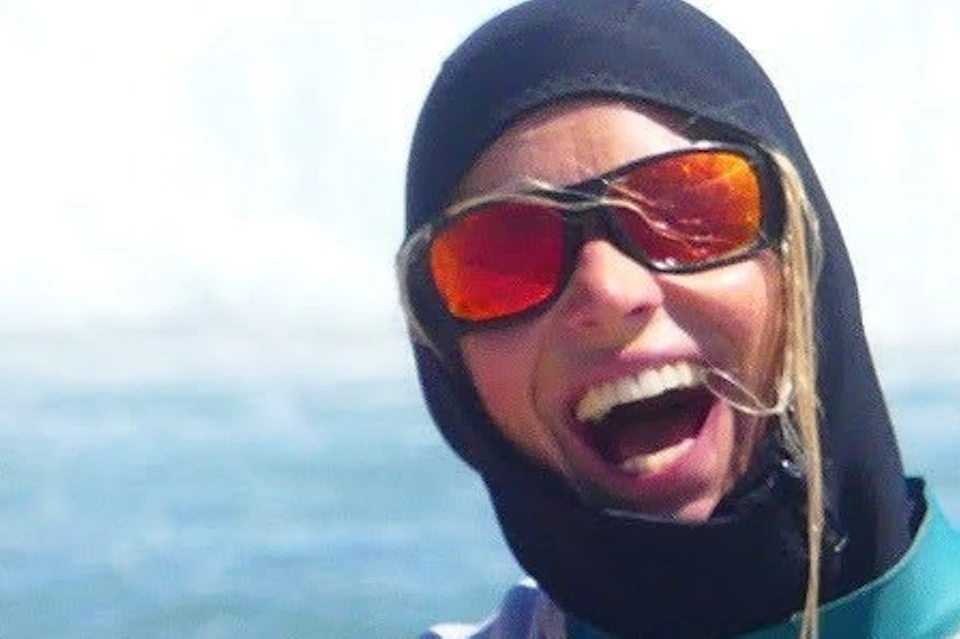 Two people in wetsuits smiling and posing with surfboards on a sunny beach, others surfing in the background.
